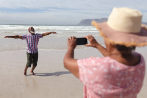 senior african american woman taking a picture of her husband on the beach. travel vacation retirement lifestyle concept